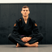 Person wearing a black martial arts uniform with orange logos sitting cross-legged on a dark background