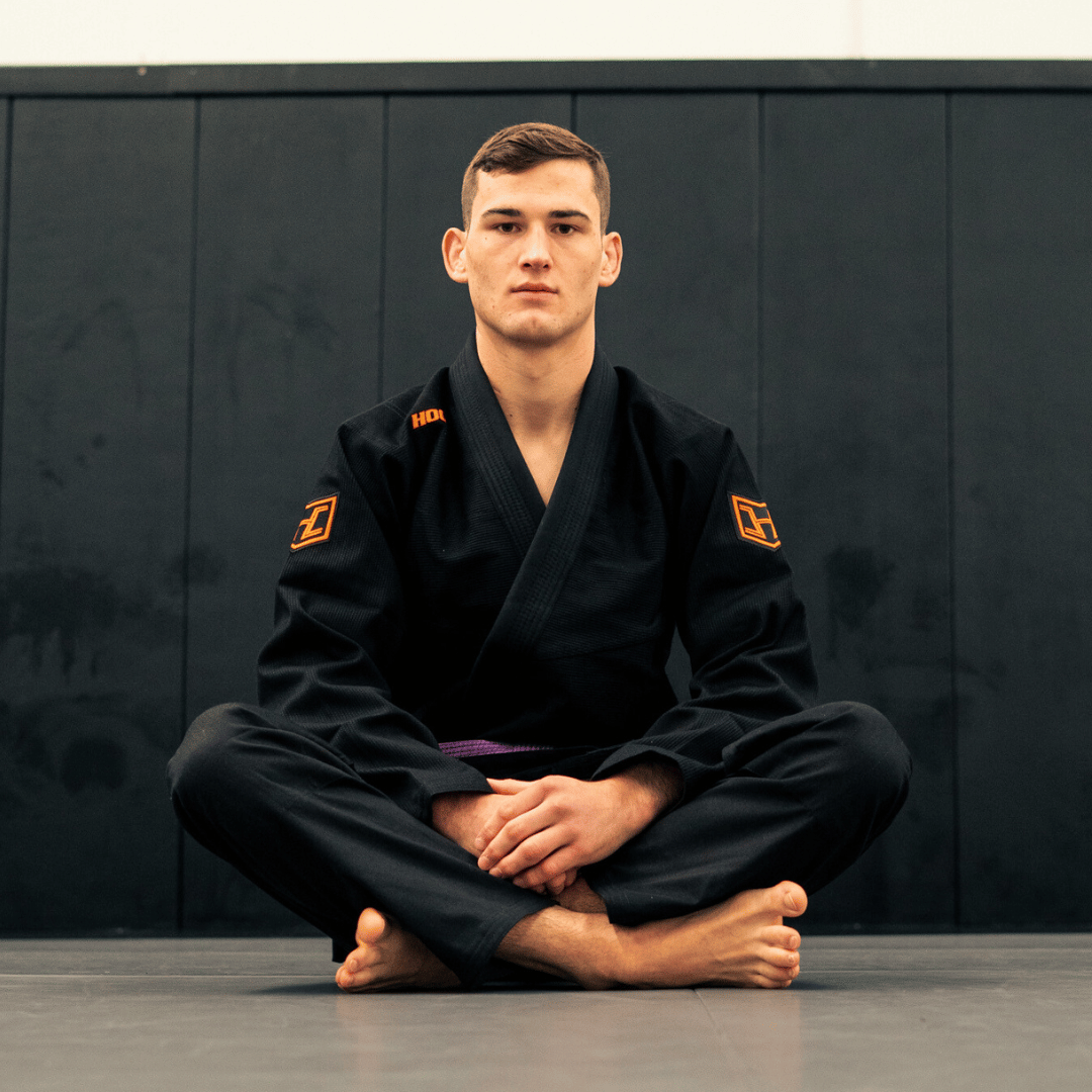 Person wearing a black martial arts uniform with orange logos sitting cross-legged on a dark background