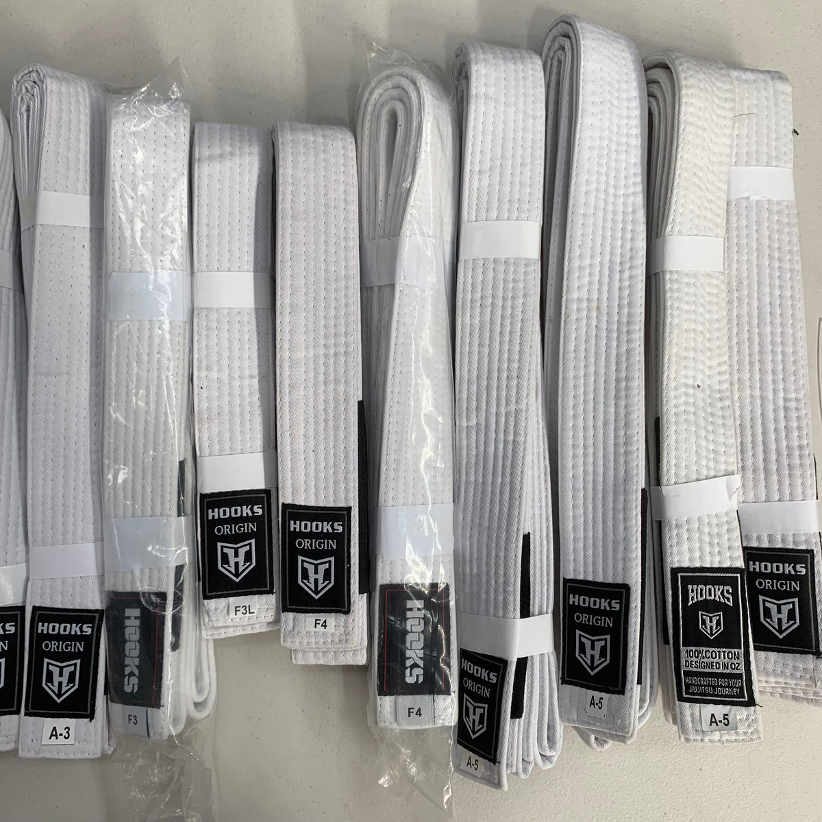 Hooks Origin White BJJ Belt, multiple folded belts in various sizes laid out showing woven texture and branded labels