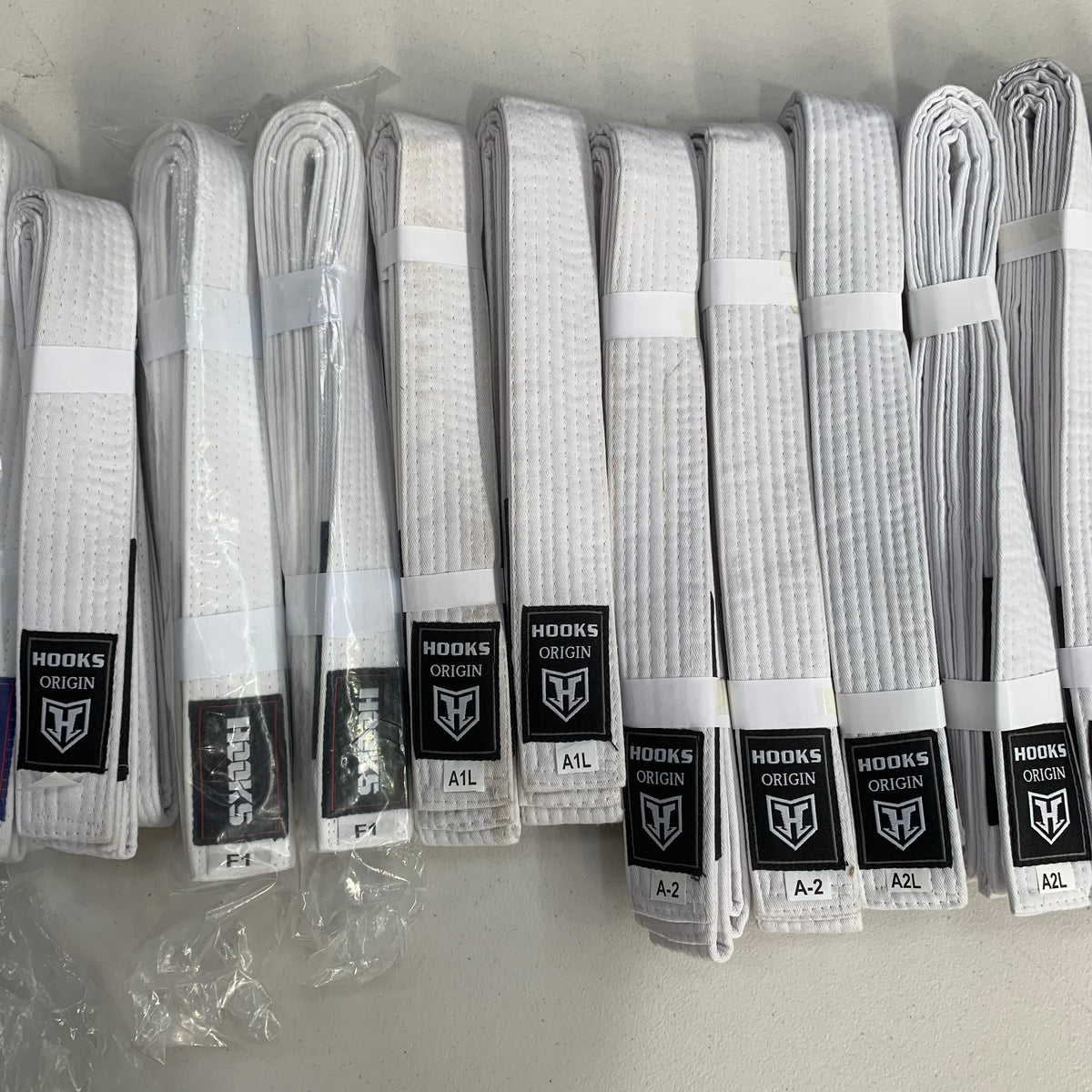 Hooks Origin White BJJ Belt, multiple folded belts laid out showing black label detail with logo patch visible.