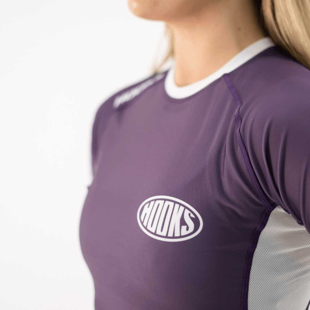 Hooks Women's Fusion Short Sleeve Rash Guard in Plum, close-up of chest logo and raglan sleeve seam, worn by a woman