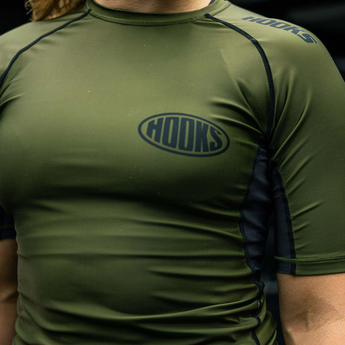 Hooks short sleeve rashguard in olive, close-up front worn view showing chest logo and black mesh side panel detail