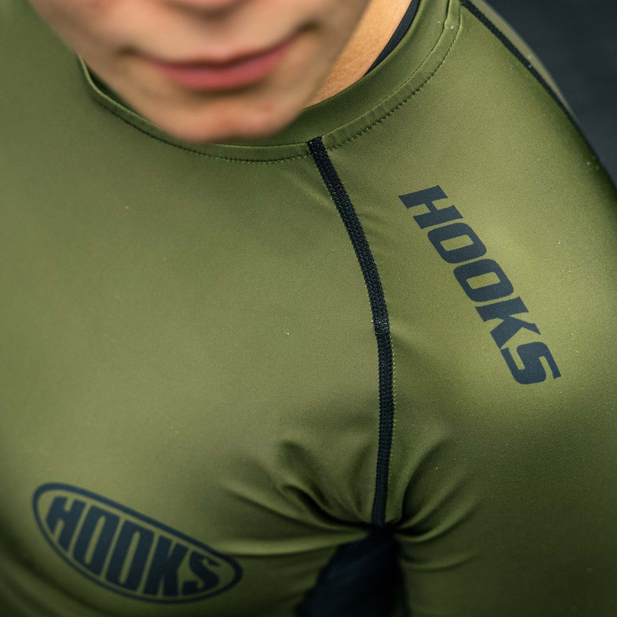 Hooks olive rashguard close-up worn, showing black chest seam stitching and shoulder logo print